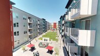 This expansive outdoor courtyard amenity space is nestled within a striking multi-story residential community, characterized by bold architectural color blocking in teal, red, and gray tones. The courtyard is designed to maximize community engagement and outdoor living, offering a diverse range of recreational and social amenities across a single, open-air level.

At the heart of the space lies a generously sized, well-manicured lawn area that provides ample room for recreation and leisure. Adjacent to the lawn, a life-size chess board with oversized game pieces serves as a unique and interactive focal point, accompanied by cornhole boards for additional outdoor entertainment.

Multiple distinct seating zones are thoughtfully arranged throughout the courtyard. A central lounge area features a built-in fire pit table surrounded by rattan sectional sofas and accent chairs adorned with vibrant teal, orange, and multicolored cushions. Additional wicker lounge groupings with teal cushions are positioned near a decorative architectural wall element, creating more intimate conversation areas.

A covered outdoor kitchen station is equipped with a stainless steel grill, bar counter, and café-style string lighting overhead, ideal for outdoor cooking and entertaining. Colorful market umbrellas in red and teal are distributed throughout the space, providing shade over additional patio dining and seating areas. Planters with ornamental trees add greenery and definition to the space, completing this well-appointed and lively communal outdoor environment.