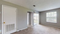 This single-story, one-bedroom, one-bathroom unit offers a straightforward and functional layout designed for comfortable everyday living. The main living area is generously proportioned, featuring neutral gray walls, plush wall-to-wall carpeting, and a textured ceiling. A sliding glass door fitted with vertical blinds allows natural light to fill the space while providing access to an outdoor area. An additional window further brightens the room, and a dedicated entryway with a front door creates a clear sense of arrival.

Adjacent to the living space, a dining area is defined by a pendant light fixture overhead, with an open passthrough connecting directly to the kitchen. The galley-style kitchen is efficiently organized, featuring white cabinetry above and below, speckled countertops, a stainless steel sink, a dishwasher, a coil-top electric range, and a full-size refrigerator. Rich wood-look flooring runs throughout the kitchen, providing a warm contrast to the lighter tones found elsewhere in the unit.

The bedroom is a comfortable, carpeted room equipped with a ceiling fan with light fixture and a window that allows natural light in. Two doors from the bedroom provide access to what appears to be a closet and a hallway, contributing to the unit's practical flow and storage options.
