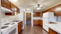 This single-story unit is designed with practicality and comfort in mind. The kitchen serves as a central feature, showcasing warm medium-toned wood cabinetry that lines both walls, providing generous storage space. Light-colored countertops offer ample prep area, while white appliances—including a range, refrigerator, and dishwasher—keep the space cohesive. Dark hardwood-style flooring runs throughout the kitchen and entry areas, adding a grounding contrast to the lighter walls and cabinetry. A ceiling fan with integrated lighting is centrally positioned, contributing to both air circulation and illumination. The entry area includes a mirrored closet alongside a solid wood front door, creating a practical transition zone into the unit. The bedroom is carpeted in a neutral tone, offering a soft underfoot feel, and benefits from natural light through a window fitted with vertical blinds. A ceiling fan keeps the room comfortable year-round, and a baseboard heater provides supplemental warmth. The bathroom is finished with white square tile surrounding a full tub-shower combination, complemented by tile flooring, a wood-toned vanity with a white countertop, and chrome fixtures—delivering a clean, straightforward aesthetic throughout.