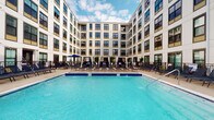 Located at 265 E State St in the heart of Columbus, Ohio, this amenity space centers around a striking resort-style outdoor pool set within a landscaped courtyard. The pool area is framed on all sides by a modern multi-story building with a clean, contemporary façade featuring large windows, a neutral white and beige exterior, and bold dark window framing that lends a sophisticated urban aesthetic. The expansive pool is finished with vivid blue tile work and is complemented by a generously sized surrounding deck. An array of lounge chairs lines the poolside, offering ample space for relaxation, while private cabanas and market umbrellas provide shaded retreats. Lush potted greenery adds a touch of natural warmth to the hardscape setting. The courtyard's enclosed layout creates a sense of privacy while still opening up to the open sky above, allowing for abundant natural light throughout the day. The overall ambiance is polished and resort-inspired, offering a refined outdoor retreat within a dynamic urban environment in downtown Columbus.