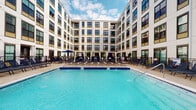 Located at 265 E State St in the heart of Columbus, Ohio, this amenity space centers around a striking resort-style outdoor pool set within a landscaped courtyard. The pool area is framed on all sides by a modern multi-story building with a clean, contemporary façade featuring large windows, a neutral white and beige exterior, and bold dark window framing that lends a sophisticated urban aesthetic. The expansive pool is finished with vivid blue tile work and is complemented by a generously sized surrounding deck. An array of lounge chairs lines the poolside, offering ample space for relaxation, while private cabanas and market umbrellas provide shaded retreats. Lush potted greenery adds a touch of natural warmth to the hardscape setting. The courtyard's enclosed layout creates a sense of privacy while still opening up to the open sky above, allowing for abundant natural light throughout the day. The overall ambiance is polished and resort-inspired, offering a refined outdoor retreat within a dynamic urban environment in downtown Columbus.