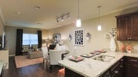 This single-story space showcases an open-concept design that integrates the kitchen, dining, and living areas into one cohesive, flowing layout. The kitchen is anchored by a substantial granite countertop with distinctive veining, functioning as both a workspace and casual bar seating area. A deep stainless steel sink and pendant lighting — featuring two hanging fixtures with clean cylindrical shades — add a polished, contemporary touch overhead.

The dining area sits adjacent to the kitchen and features a round dark-wood table surrounded by upholstered chairs in a bold chevron pattern, providing a striking visual contrast against the warm, neutral walls. A coordinating set of place settings with coral accents adds a curated, styled touch to the space.

The living room, visible beyond the dining area, is warm and inviting, with a sectional sofa, area rug in coral tones, and layered lighting from a floor lamp and natural light filtering through curtained windows. Track lighting runs along the ceiling, providing versatile illumination throughout. Dark hardwood-style flooring grounds the entire space, complementing the overall warm, modern aesthetic. Decorative wall art and mirrors enhance the visual depth throughout.