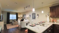 This single-story space showcases an open-concept design that integrates the kitchen, dining, and living areas into one cohesive, flowing layout. The kitchen is anchored by a substantial granite countertop with distinctive veining, functioning as both a workspace and casual bar seating area. A deep stainless steel sink and pendant lighting — featuring two hanging fixtures with clean cylindrical shades — add a polished, contemporary touch overhead.

The dining area sits adjacent to the kitchen and features a round dark-wood table surrounded by upholstered chairs in a bold chevron pattern, providing a striking visual contrast against the warm, neutral walls. A coordinating set of place settings with coral accents adds a curated, styled touch to the space.

The living room, visible beyond the dining area, is warm and inviting, with a sectional sofa, area rug in coral tones, and layered lighting from a floor lamp and natural light filtering through curtained windows. Track lighting runs along the ceiling, providing versatile illumination throughout. Dark hardwood-style flooring grounds the entire space, complementing the overall warm, modern aesthetic. Decorative wall art and mirrors enhance the visual depth throughout.