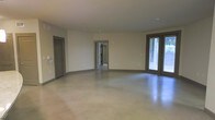 This single-floor space presents a clean, minimalist aesthetic built around an open floor plan. The most prominent feature is the polished concrete flooring, which spans the entire main area and lends a smooth, industrial character while reflecting ambient light throughout the room. The neutral, light-toned walls complement the flooring and contribute to a bright, airy atmosphere.

A set of large glass-paned double doors along one wall allows abundant natural light to flood the interior, offering views of the surrounding greenery outside. These doors also provide direct outdoor access, enhancing connectivity between the interior and exterior environments.

The layout includes multiple doorways leading to adjoining rooms, suggesting additional functional spaces beyond the main open area. Gray-trimmed interior doors provide subtle contrast against the neutral walls, adding a cohesive design detail throughout. Recessed ceiling lighting supplements the natural light, ensuring the space remains well-illuminated.

Overall, the space conveys a versatile, move-in-ready environment with a modern yet understated design sensibility, suitable for a variety of uses and configurations.