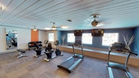 This fitness room is thoughtfully designed to support a well-rounded workout experience. The space is finished with a distinctive white shiplap ceiling, lending a clean and airy aesthetic that is complemented by ceiling fans to maintain comfortable airflow during exercise. The walls feature a bold color palette, with a vibrant orange accent wall on one side and cool blue tones on the other, creating an energizing atmosphere.

Natural light enters the room through several windows positioned along the perimeter, each framed by neutral roller shades. Flat-screen televisions are mounted above the windows, providing entertainment options for users during their workouts. The flooring consists of low-pile carpet tiles in a neutral tone, offering both durability and cushioning underfoot.

The equipment lineup includes a treadmill and a stationary recumbent bike for cardiovascular training, as well as a rack of free weights positioned along the orange accent wall for strength conditioning. Overhead, multiple ceiling-mounted light fixtures ensure the room is well-lit throughout. Overall, the space strikes a balance between functional fitness amenities and an inviting, modern design that encourages regular use.