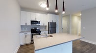 Located in the heart of Columbus, Ohio, this single-floor residence at 223 E Town St offers a thoughtfully designed living space with modern finishes throughout. The kitchen serves as a central highlight, showcasing a large quartz-topped island with an undermount sink — ideal for both meal preparation and casual dining. Crisp white shaker cabinets provide ample storage, while stainless steel appliances, including a refrigerator, electric range, and over-the-range microwave, add a polished, contemporary feel. Stylish dark pendant lights hang above the island, complementing the flush-mount ceiling fixture and creating a warm, layered lighting atmosphere. The kitchen flows seamlessly into the adjoining living areas, supported by durable wood-style flooring that extends throughout the space. Light-toned walls and bright lighting contribute to an open, airy ambiance. A hallway visible from the kitchen leads to additional areas of the unit, suggesting a well-organized and practical floor plan. Overall, this space combines functional design with modern aesthetics, making it a comfortable and attractive offering in downtown Columbus.