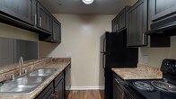 Located at 1325 Riverside Pkwy in Austell, Georgia, this single-floor residence offers a well-organized layout designed for comfortable everyday living. The open living and dining area features warm beige walls, plush carpeting, and a textured ceiling, with a pendant light fixture marking the transition between the two spaces. Hardwood-style flooring flows into the kitchen, which is laid out in a classic galley configuration. Dark espresso cabinetry with brushed hardware lines both sides of the kitchen, complemented by granite-style countertops, a double stainless steel sink, a dishwasher, and black appliances including a refrigerator and electric range. The bedrooms are generously sized, each featuring soft carpeting, neutral-toned walls, and natural light from windows with blinds. One bedroom includes access to a dedicated laundry area with hookups and built-in shelving for additional storage. Both bathrooms are appointed with dark vanity cabinetry, granite-style countertops, framed mirrors, and a tub-shower combination set over hardwood-style flooring. Throughout the residence, consistent neutral tones and practical finishes create a cohesive, move-in-ready environment suitable for a range of lifestyles.
