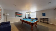 Located at 5039 Chalet Ct in Tampa, FL, this single-floor recreational space is designed with entertainment and relaxation in mind. The centerpiece of the room is a full-size billiards table with a vivid teal felt surface and a classic hardwood frame, positioned beneath a sleek, industrial-style pendant light fixture with multiple drum shades that provides excellent task lighting over the playing surface. The room is finished with neutral-toned walls accented by white chair rail molding, and carpeted flooring in a subtle woven pattern that adds texture underfoot. Two vibrant large-scale abstract paintings adorn the walls, introducing bold splashes of color that energize the space. A wall-mounted cue rack keeps billiards accessories neatly organized and easily accessible. A cozy seating area near the windows features plush navy blue accent chairs paired with a gold geometric side table, creating a comfortable retreat for spectators. Multiple windows fitted with white horizontal blinds allow natural light to filter in while maintaining privacy. A bar-height table and stools provide an additional casual gathering spot. A staircase with wood-look flooring and a pass-through interior window connects to an adjacent area, offering views into what appears to be a front office or reception space. Overall, the room strikes a balance between functional recreation and polished design.