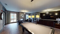 Located at 4380 Eastgate Blvd in Cincinnati, OH, this single-floor community clubhouse offers a sophisticated and well-appointed gathering space designed for both relaxation and recreation. The layout flows seamlessly between distinct areas, each finished with dark wood trim, patterned tile flooring, and warm, layered lighting throughout.

The central game room is anchored by a full-size billiards table beneath stylish pendant lighting, with open sightlines connecting to the lounge and entry areas. Adjacent to the game room, a dedicated card room features a round poker table surrounded by cream upholstered chairs, set beneath a striking coffered ceiling with teal accents and recessed lighting, with a large window bringing in natural light.

The lounge area is a standout, centered on a floor-to-ceiling stacked stone fireplace framed by elegant drapery and sliding glass doors that open to an outdoor patio. Leather seating and an orb chandelier contribute to the refined atmosphere. A media wall with dark wood paneling, built-in sconces, and a wall-mounted television provides a dedicated entertainment area with comfortable seating nearby.

The full catering kitchen is equipped with dark cabinetry, granite countertops, a red tile backsplash, stainless steel appliances, and a center island, making it ideal for events and gatherings. A fitness center is also accessible directly from the clubhouse.