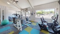 This fitness center is housed in a single-story structure characterized by vaulted, beam-accented ceilings and white shiplap walls that lend the space a clean, airy aesthetic. Large windows line the perimeter, flooding the interior with abundant natural light and offering views of the surrounding outdoor greenery. The flooring features a vibrant patchwork of teal, yellow-green, and gray carpet tiles, adding a modern, energetic feel to the space. The facility is comprehensively equipped for a full-body workout, including True-brand treadmills, a stationary spin bike, an Expresso recumbent bike, and an elliptical machine for cardio training. A multi-station cable weight machine and a dedicated free-weight rack provide ample strength training options, while an adjustable workout bench and a stability ball round out the functional fitness offerings. A glass-paneled door opens directly to an outdoor area, reinforcing the connection to the natural surroundings and providing convenient access. Posted fitness center rules are prominently displayed near the entrance, and a ceiling fan contributes to air circulation throughout. The overall ambiance is bright, welcoming, and purpose-built to support a wide range of fitness routines.
