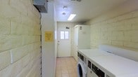 This shared laundry facility is a single-floor, purpose-built space designed for practicality and ease of use. The room is lined with painted white brick walls that give it a clean, utilitarian character, while light-toned tile flooring runs throughout, making maintenance straightforward. Overhead, a flush-mounted fluorescent light panel provides bright, even illumination across the entire space. A row of commercial-grade front-loading washers and top-loading dryers lines one wall, offering multiple machines to accommodate simultaneous use. A folding counter sits adjacent to the machines, providing a convenient surface for handling laundry. A small window beside the entry door allows for a modest amount of natural light, contributing to the room's clean, well-lit atmosphere. The entry door features a security camera mounted above it, reflecting attention to the safety of the facility. Informational signage is posted on the walls, guiding users on proper machine use and facility etiquette. A no-smoking notice is also clearly displayed, reinforcing the clean and shared nature of the environment. Overall, this laundry room is a well-organized, functional communal facility built to serve the practical needs of its users efficiently.