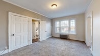 This well-appointed single-floor space blends classic character with thoughtful updates throughout. The main living area is generously sized, featuring neutral carpeting, crown molding, and a paneled entry door that sets a traditional tone. Natural light pours in through large, multi-pane windows, and a cast-iron radiator underscores the home's period charm.

The kitchen has been tastefully renovated with crisp white shaker cabinetry, granite-style countertops, a white subway tile backsplash, and wood-look flooring. Black hardware and appliances — including a gas range and a full-size refrigerator — add a modern contrast to the clean, bright palette.

The spacious bedroom continues the warm, neutral tones with plush carpeting, crown molding, and bright windows that overlook the surrounding neighborhood. Classic six-panel white doors lead to a walk-in closet area featuring built-in shelving and hanging rods across multiple sections, offering generous storage capacity on rich hardwood-style floors.

The full bathroom retains its vintage appeal with white square wall tiles accented by bold black trim details, a pedestal sink, a tub-shower combination, and a window that allows natural light to brighten the space. Wood-look flooring ties the room together with a clean, updated finish.

Overall, this space presents a cohesive blend of timeless architectural character and practical modern updates.