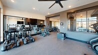 Located at 15101 Royal Creek Dr in Fuquay-Varina, NC, this well-appointed fitness center is thoughtfully designed to serve a full range of workout needs across a single open floor. The space is organized into distinct functional zones, each with its own character and equipment focus. The cardio area features treadmills, a stair climber, a rowing machine, and stationary bikes, all positioned alongside expansive floor-to-ceiling windows that flood the room with natural light and offer open views of the surrounding landscape. Large ceiling fans overhead provide airflow and comfort throughout. The strength training zone is equipped with a multi-functional cable machine system, a dedicated dumbbell rack, and a leg press machine. Full-length mirrors line the walls, visually expanding the space while supporting proper form. A separate studio room with light wood-look flooring, mirrored walls, a ballet barre, and cycling bikes provides a clean, versatile space for group fitness or focused training sessions. The design palette features a sophisticated contrast of dark charcoal accent walls, crisp white ceilings, and warm wood flooring, complemented by modern wall sconces and recessed lighting throughout. The overall ambiance is sleek, functional, and contemporary, creating an inviting environment for all levels of fitness.