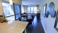 This single-floor residence showcases an open-concept design that integrates the dining and living areas into one cohesive, flowing space. Dark hardwood flooring runs throughout, providing a rich, grounding contrast to the bright, airy atmosphere created by expansive floor-to-ceiling windows along multiple walls. These generous windows frame striking urban skyline views, flooding the interior with natural light and reinforcing the elevated city setting.

The dining area features a round glass-top table paired with sleek, high-backed navy blue chairs, positioned adjacent to a marble-topped kitchen counter that defines the boundary between the kitchen and the open living space. The living area is comfortably appointed with a sofa, accent chairs, and a coffee table arranged to take full advantage of the panoramic city backdrop. Neutral wall tones and white trim complement the darker flooring, while carefully placed lamps and framed artwork add warmth and a curated aesthetic. The overall ambiance is sophisticated and contemporary, balancing urban energy with a refined residential feel.