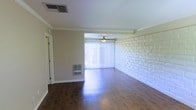 This single-floor space offers a clean and functional layout with a series of distinctive architectural details that give it notable character. The main living area is anchored by rich, dark hardwood flooring that reflects light beautifully, adding warmth and depth to the interior. A standout feature is the full-height painted brick accent wall, which adds texture and a classic structural element to the space, contrasting pleasantly with the neutral-toned walls and crisp white crown molding that lines the ceiling throughout.

The living area benefits from an abundance of natural light, streaming in through large sliding glass doors dressed with vertical blinds, which also provide access to the outdoors. A ceiling fan is centrally positioned in this area, ensuring year-round comfort and air circulation. The space also includes a wall-mounted heating unit, highlighting practical functionality alongside its aesthetic appeal.

Crown molding trims the ceilings throughout, adding a refined architectural finish, while the ceiling vents and clean baseboards speak to a well-maintained interior. The open flow between areas creates a sense of spaciousness, making the layout feel both airy and versatile. Overall, the space presents a blend of classic charm and everyday practicality.