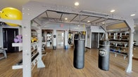 This fitness center is thoughtfully designed to support a wide range of workout styles, from high-intensity combat training to cardio and strength conditioning. The space is organized into distinct functional zones, each outfitted with purpose-built equipment to maximize usability.

The centerpiece of the facility is a dedicated combat and functional training area featuring multiple heavy punching bags suspended from a robust overhead rig. The same rig supports TRX suspension trainers, enabling bodyweight and resistance-based exercises. White open-frame shelving units line the walls, neatly storing accessories such as medicine balls, kettlebells, and stability balls, keeping the floor space open and uncluttered.

Adjacent to the training area, a strength zone houses a cable machine, adjustable bench, and additional resistance equipment, all positioned on the same warm hardwood-style flooring that runs throughout the space. Recessed lighting provides consistent, even illumination across the entire floor.

A separate cardio section features treadmills, an elliptical trainer, a stair-stepper, and a rowing machine, arranged in front of a large window that admits natural light and exterior views. The overall ambiance is clean, modern, and well-organized, with neutral gray walls contributing to a focused and functional training environment.