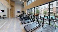 This well-appointed fitness center offers a sophisticated and comprehensive workout environment designed to meet a wide range of exercise needs. The space spans two levels, with the upper mezzanine level featuring a sleek railing and overlooking the main fitness floor below, creating an open, airy atmosphere with impressively soaring ceilings.

Large, floor-to-ceiling windows line the perimeter of the main floor, flooding the space with abundant natural light and offering views of an outdoor courtyard and pool area. The cardio zone is generously equipped with a row of treadmills, elliptical machines, and stair climbers, all positioned to take advantage of the outdoor views. A dedicated strength training area features a cable machine, Smith machine, weight bench, and a full dumbbell rack, catering to a variety of lifting routines.

A separate functional training nook houses stability balls, medicine balls, yoga mats, a heavy bag, and additional benches, providing a versatile space for flexibility and cross-training workouts. Wall-mounted televisions are positioned throughout for entertainment during exercise.

The interior design is elevated with herringbone wood-paneled accent walls, large floor mirrors that enhance the sense of space, patterned carpet flooring, and warm ambient lighting. A stylish entryway with geometric pendant lighting and cushioned seating completes the polished, upscale aesthetic of this fitness facility.