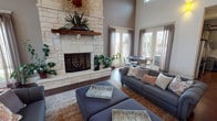 This expansive single-floor clubhouse is designed to accommodate a wide range of social and recreational activities. The main gathering area commands attention with soaring ceilings and a dramatic floor-to-ceiling limestone fireplace with a rustic wood mantle, anchoring a comfortable lounge furnished with plush gray sofas and ottomans. Abundant natural light pours through multiple sets of French doors adorned with sheer gray drapery, offering views of the outdoor pool and surrounding community.

The space flows into several distinct activity zones, including a billiards area with a full-size pool table, a putting green, and intimate leather seating nooks decorated with eclectic wall art and trailing greenery. High pub-style tables with uniquely styled stools are positioned near the French doors, creating casual spots for conversation.

The fully equipped kitchen is a standout feature, showcasing rich olive-green cabinetry, granite countertops, a large center island with barrel-style seating, stainless steel appliances, and a bold stainless hood vent — ideal for community gatherings and catered events. Dark hardwood flooring runs throughout, tying together the warm, rustic-modern aesthetic that defines this vibrant and versatile clubhouse.