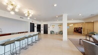 Located at 10 Commerce Dr in New Rochelle, NY, this contemporary amenity lounge is thoughtfully designed to provide a sophisticated and comfortable communal environment. The single-level space is defined by clean architectural lines, white walls, and large-format light tile flooring that flows seamlessly throughout.

The main lounge area is anchored by a curated seating arrangement of modern sofas and accent chairs, complemented by vibrant teal upholstered ottomans that add a bold pop of color. Floor-to-ceiling glass windows and sliding doors open the space to an outdoor terrace, flooding the interior with abundant natural light and offering direct views of the landscaped exterior amenity area.

A sleek kitchenette and bar counter serves as a central social hub, featuring warm wood cabinetry, a white countertop with bar seating, and a statement sputnik chandelier overhead. Adjacent to the main lounge, a secondary seating alcove offers a quieter setting with sage green armchairs, wall sconces with gold accents, and curated artwork — ideal for informal meetings or relaxed conversation.

A wall-mounted television and additional lounge seating create a media-friendly zone, while the open layout and structural columns provide a sense of spaciousness. The overall ambiance is modern, welcoming, and refined, making this space well-suited for both social gatherings and quiet relaxation.
