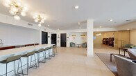 Located at 10 Commerce Dr in New Rochelle, NY, this contemporary amenity lounge is thoughtfully designed to provide a sophisticated and comfortable communal environment. The single-level space is defined by clean architectural lines, white walls, and large-format light tile flooring that flows seamlessly throughout.

The main lounge area is anchored by a curated seating arrangement of modern sofas and accent chairs, complemented by vibrant teal upholstered ottomans that add a bold pop of color. Floor-to-ceiling glass windows and sliding doors open the space to an outdoor terrace, flooding the interior with abundant natural light and offering direct views of the landscaped exterior amenity area.

A sleek kitchenette and bar counter serves as a central social hub, featuring warm wood cabinetry, a white countertop with bar seating, and a statement sputnik chandelier overhead. Adjacent to the main lounge, a secondary seating alcove offers a quieter setting with sage green armchairs, wall sconces with gold accents, and curated artwork — ideal for informal meetings or relaxed conversation.

A wall-mounted television and additional lounge seating create a media-friendly zone, while the open layout and structural columns provide a sense of spaciousness. The overall ambiance is modern, welcoming, and refined, making this space well-suited for both social gatherings and quiet relaxation.