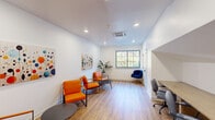 This single-floor office space offers a clean, modern aesthetic designed for professional functionality. The room features light wood-tone flooring throughout, crisp white walls, and a ceiling equipped with recessed lighting and a central HVAC unit, ensuring a well-lit and climate-controlled environment.

The seating area is furnished with bold orange chairs arranged around low, round side tables, creating a welcoming and visually dynamic lounge or waiting area. A navy blue upholstered task chair adds contrast and variety. Two vibrant abstract artworks depicting colorful circles and dots are prominently displayed on the walls, lending the space an energetic, contemporary character.

A built-in wood-surface desk runs along one wall, accompanied by gray ergonomic office chairs — providing a dedicated workstation zone. A glass-paned door leads to an adjacent corridor, and a window on one wall allows natural light to filter in with views of greenery outside, adding a refreshing connection to the outdoors.

A potted tropical plant near the window further softens the space and enhances its lively ambiance. The layout efficiently combines a seating/reception area with a functional workspace, making it a well-appointed and versatile office environment.