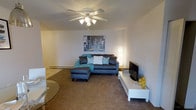 This single-floor residence offers a comfortable and thoughtfully arranged living space designed for everyday functionality. The living room serves as the heart of the home, featuring wall-to-wall carpeting that adds warmth underfoot. A ceiling fan with integrated lighting is centrally mounted, providing both air circulation and ambient illumination. The room accommodates a sectional sofa accented with teal and white throw pillows, complemented by a soft area rug that defines the seating zone. A low-profile white media console houses the television and provides additional storage, while a sleek floor lamp in the corner contributes to the room's layered lighting. Neutral-toned walls offer a clean, versatile backdrop, and a baseboard heating unit runs along the wall, ensuring year-round comfort. The space has an open, airy feel with a straightforward layout that maximizes usability. Decorative wall art adds a touch of personality, and subtle accent pieces throughout contribute to the cohesive, modern aesthetic. Overall, this residence presents a practical and welcoming environment suited for comfortable single-floor living.