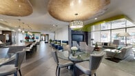 This expansive single-floor amenity space is a showcase of contemporary design, blending bold colors, high-end finishes, and thoughtfully arranged functional zones. The entrance welcomes visitors with large-format tile flooring, lush potted greenery, and sleek black glass doors that set a polished tone throughout.

The main lounge area is defined by striking chartreuse-lacquered ceiling panels that contrast beautifully against crisp white walls, while a sculptural drum pendant and recessed lighting illuminate the space. Multiple seating arrangements — including plush gray recliners, blue leather lounge chairs with lime-green accent pillows, and glass-top dining tables with upholstered chairs — create distinct areas for relaxation and socializing. A wall-mounted flat-screen television and a teal accent wall anchor one of the lounge corners.

A standout feature is the open game room, centered around a modern pool table beneath warm pendant lighting, with floor-to-ceiling sheer curtains filtering abundant natural light from large exterior windows. Textured wave-pattern wall panels add architectural depth.

The gourmet kitchen is a true centerpiece, outfitted with an expansive granite waterfall island, sleek dark cabinetry, stainless steel appliances including a professional-grade refrigerator and double wall ovens, and a series of elegant crystal pendant lights — the perfect space for entertaining.