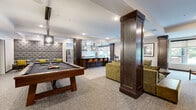 This thoughtfully designed single-floor amenity lounge seamlessly blends recreation, socializing, and comfort within one open, flowing layout. The space is anchored by a generously sized billiards table positioned beneath pendant lighting, with cue racks mounted on the wall nearby and framed artwork adding a refined touch. Dark-stained wooden columns punctuate the open floor plan, providing subtle zone definition while reinforcing the space's sophisticated character.

The fully equipped kitchen is a standout feature, outfitted with stainless steel appliances including a refrigerator, dishwasher, and range with a wall-mounted hood vent. White subway tile backsplash, open shelving, and a large dark granite-topped island with leather bar stools create an inviting and functional gathering hub. Geometric patterned tile flooring grounds the kitchen area, while large windows along the perimeter flood the space with natural light and offer views of lush greenery outdoors.

Adjacent to the kitchen, a comfortable lounge area features plush olive-green and leather seating arranged around an organic glass-topped coffee table, fostering relaxed conversation. A tufted gray accent wall panel adds a bold textural element near the seating nook. Double glass-paned entry doors with recessed tray ceilings above complete the polished, welcoming atmosphere throughout.