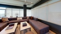 Located at 222 M St SW in Washington, DC, this single-floor communal space showcases a thoughtfully designed interior that balances modern aesthetics with functional living. The open layout connects multiple distinct zones, creating a cohesive environment ideal for socializing and relaxation.

The lounge area features a generous tufted sectional sofa in rich brown upholstery, arranged around a sleek white coffee table. Large windows fitted with roller shades allow diffused natural light to fill the room, complemented by recessed ceiling lighting. Deep charcoal accent walls add a sophisticated contrast throughout.

The kitchen and social hub area is anchored by a substantial island with warm walnut wood cabinetry and a crisp white countertop, accompanied by contemporary bar seating. Pendant lighting overhead and light hardwood flooring contribute to a refined yet inviting atmosphere. A wall-mounted television and tiled backsplash enhance the kitchen's functionality and style.

Bold, large-format photographic murals depicting vibrant cityscapes and atmospheric light scenes serve as striking focal points throughout the space, infusing energy and visual interest. Additional bar-height seating and open sightlines to exterior windows reinforce the space's social, community-oriented character.