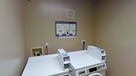 This single-floor laundry facility is a dedicated, functional space designed to serve shared-use needs efficiently. The room features a clean, neutral color palette with beige walls that contribute to a bright and utilitarian ambiance. At the center of the space, a row of modern front-loading washing machines and dryers are neatly arranged side by side, each unit topped with a flat surface that provides convenient space for folding or staging laundry. The machines appear to be operated via digital controls, and a clearly posted operating instructions placard is mounted on the wall above the units, offering guidance on both washer and dryer use — a practical and user-friendly touch. The room is equipped with standard electrical outlets along the walls to support the appliances, and a utility access panel is visible near the floor on one side. Ventilation is provided by a ceiling vent positioned above the machines, ensuring proper airflow throughout the space. The overall layout is straightforward and well-organized, prioritizing ease of use and accessibility. The space is well-maintained, with clean flooring and walls, reflecting a tidy and orderly environment suited for communal laundry use.