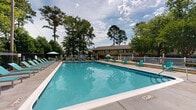Located at 5034 Hunt Club Rd in Wilmington, North Carolina, this outdoor amenity space centers around a generously sized swimming pool that serves as the focal point of the area. The pool is enclosed by a sturdy wrought-iron fence, ensuring both safety and security, with a concrete pool deck that provides ample space for lounging and relaxation. Numerous teal-colored Adirondack chairs and chaise lounges are arranged around the perimeter, offering plenty of seating for sunbathing and socializing. A shaded umbrella station provides a comfortable retreat from the North Carolina sun. An elevated wooden deck with a white railing adjoins one side of the pool area, adding a distinct gathering space complete with additional seating. The surrounding landscape is thoughtfully maintained, featuring well-kept green lawns, ornamental plantings, decorative rock beds, and towering mature pine and deciduous trees that lend a sense of privacy and natural beauty to the space. A paved walkway connects the pool enclosure to the broader grounds, making the area easily accessible. The combination of open sky views, verdant tree canopy, and carefully curated outdoor furnishings creates a welcoming and resort-like atmosphere ideal for outdoor recreation and leisure.