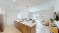 This well-appointed single-floor residence showcases a contemporary design with a bright, open-concept layout. The kitchen serves as the centerpiece of the living space, anchored by a generously sized island topped with white quartz countertops and warm wood cabinetry with gold-toned hardware. Pendant lighting hangs above the island, which offers bar-style seating for casual dining. The kitchen is fully equipped with stainless steel appliances, including a range, microwave, refrigerator, and dishwasher, complemented by crisp white upper cabinets and light hardwood-style flooring that flows throughout the main living areas. The open living area extends naturally from the kitchen, featuring a cozy seating nook with natural light from large windows fitted with horizontal blinds. Two bedrooms offer a calm retreat, each carpeted and illuminated by natural light through windows with outdoor views. The bathroom is finished with warm wood vanity cabinetry, granite countertops, an illuminated LED mirror, and a glass-enclosed walk-in shower with large-format tile surround. Throughout the residence, a neutral, light-filled palette creates a clean and cohesive modern ambiance.