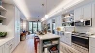 This thoughtfully designed 1-bedroom, 1-bathroom residence offers an efficient yet stylish layout across a single floor. The open-concept kitchen is a standout, featuring light gray shaker cabinetry with brass hardware, white quartz countertops, stainless steel appliances, open shelving, and a two-tone kitchen island with seating — all illuminated by globe pendant lights suspended from a tray ceiling. Light hardwood flooring flows throughout, connecting the kitchen to a built-in desk nook and seamlessly into the living area. The living room is bright and welcoming, with floor-to-ceiling glass doors opening to a private balcony, allowing abundant natural light and outdoor views. A grid-style mirror accent wall adds depth and character. The bedroom is comfortably sized with large windows that frame outdoor scenery, complemented by two closets for generous storage. The bathroom features large-format tile flooring, a marble-tiled walk-in shower with a rainfall showerhead, and gray vanity cabinetry with brass fixtures. Throughout the residence, high-end finishes, cohesive modern design, and carefully considered details create a polished and functional living environment.