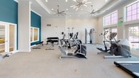 This fitness center in Canton, Michigan offers a spacious, single-floor layout designed to accommodate a comprehensive workout experience. The interior is finished with a clean, modern aesthetic, featuring crisp white walls accented by bold teal feature walls that add a dynamic visual element to the space. The ceiling is notably elevated, enhanced by a tray ceiling detail and fitted with a combination of recessed lighting and stylish sputnik-style chandeliers, along with large ceiling fans that help maintain air circulation throughout the room.

Natural light fills the space generously through large, multi-pane windows, particularly in the cardio section, where a row of True treadmills lines the wall beneath floor-to-ceiling windows overlooking the outdoors. Additional cardio equipment includes multiple Cybex elliptical trainers and stationary bikes, providing ample options for aerobic training.

The strength training area features a cable and pulley multi-station machine, a vertical knee raise tower, and a dedicated free weight section with a tiered dumbbell rack offering a full range of weights accompanied by a flat utility bench. The flooring throughout is a speckled, durable surface suited for high-traffic fitness use. Overall, the space presents a bright, well-organized, and inviting environment for a full range of fitness activities.