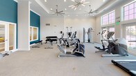 This fitness center in Canton, Michigan offers a spacious, single-floor layout designed to accommodate a comprehensive workout experience. The interior is finished with a clean, modern aesthetic, featuring crisp white walls accented by bold teal feature walls that add a dynamic visual element to the space. The ceiling is notably elevated, enhanced by a tray ceiling detail and fitted with a combination of recessed lighting and stylish sputnik-style chandeliers, along with large ceiling fans that help maintain air circulation throughout the room.

Natural light fills the space generously through large, multi-pane windows, particularly in the cardio section, where a row of True treadmills lines the wall beneath floor-to-ceiling windows overlooking the outdoors. Additional cardio equipment includes multiple Cybex elliptical trainers and stationary bikes, providing ample options for aerobic training.

The strength training area features a cable and pulley multi-station machine, a vertical knee raise tower, and a dedicated free weight section with a tiered dumbbell rack offering a full range of weights accompanied by a flat utility bench. The flooring throughout is a speckled, durable surface suited for high-traffic fitness use. Overall, the space presents a bright, well-organized, and inviting environment for a full range of fitness activities.