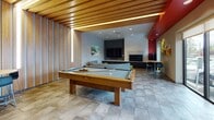 Located at 940 North Ruby Street in Spokane, Washington, this single-floor community amenity space is designed to encourage social interaction and relaxation. The interior is immediately striking, with a bold design palette that combines warm wood slat ceiling accents, vivid red architectural details, and large floor-to-ceiling windows that flood the space with natural light and offer clear views of the surrounding streetscape.

The main lounge area anchors the space with a generous curved sectional sofa upholstered in a patterned fabric, paired with a round wood coffee table and bright chartreuse ottomans that add a playful pop of color. A mounted flat-screen television and a textured stone fireplace surround create a comfortable media and gathering focal point. Adjacent to the lounge, a full-size billiard table set beneath the warm glow of the wood-paneled ceiling invites friendly competition.

A café-style high-top table and bar seating near the windows provide a casual spot for working or socializing, while a pair of upholstered barrel chairs near the entrance offer a quieter seating option. Colorful contemporary artwork is displayed throughout, reinforcing the modern, energetic atmosphere. Stone-look tile flooring ties the entire space together, balancing the warmth of the wood ceiling elements with a clean, durable finish suited for high-traffic use.