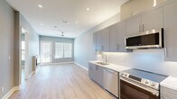 Located at 1911 Park Ave in Dallas, TX, this thoughtfully designed one-bedroom, two-bathroom residence offers a clean, contemporary aesthetic throughout. Light wood flooring flows seamlessly across the open-concept living and kitchen area, creating a bright and cohesive atmosphere. The modern kitchen is a standout feature, outfitted with sleek grey cabinetry, a white penny-tile backsplash, white quartz countertops, and a full suite of stainless steel appliances including a side-by-side refrigerator, electric range, built-in microwave, and dishwasher. A ceiling fan and recessed lighting enhance the comfort of the living area, which is bathed in natural light from large windows overlooking the outdoors. The bedroom is a well-proportioned retreat with its own window, ceiling fan, and access to a walk-in closet featuring built-in shelving and hanging rods. A stacked washer and dryer unit is conveniently tucked within a dedicated closet near the entryway. Cool grey walls with crisp white trim lend a clean, modern feel throughout. Residents also have access to an elevated rooftop terrace with panoramic views of the Dallas skyline and surrounding treetops, offering a compelling outdoor extension of the living space.