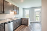 This single-floor space offers a well-designed layout with a clean, modern aesthetic carried consistently throughout. The kitchen serves as a functional centerpiece, outfitted with dark-toned cabinetry, granite countertops, and a full suite of stainless steel appliances including a refrigerator with ice maker, electric range, built-in microwave, and dishwasher. A peninsula provides additional counter space and utility, while recessed lighting illuminates the area effectively. Light-toned wood-look flooring extends throughout the space, contributing to a cohesive and contemporary feel.

The living area is spacious and open, featuring a ceiling fan with light fixture and a wall-mounted pass-through window that connects to an adjacent space. Natural light enters generously through windows fitted with blinds, and a glass exterior door offers views of the surrounding outdoor landscape. A wall-unit HVAC system provides climate control.

The bathroom is finished with a granite-topped vanity featuring dark wood cabinetry and drawer storage, a large frameless mirror, and a tub-shower combination. Neutral gray walls and tiled flooring complete the polished, practical design of the room.