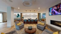 Located at 211 Concord Turnpike in Cambridge, Massachusetts, this single-floor amenity space is designed with a contemporary aesthetic that balances social gathering and leisure. The open layout is anchored by a central lounge area featuring plush upholstered armchairs arranged around a warm-toned circular coffee table, all set atop a patterned area rug that adds visual depth to the light wood flooring running throughout.

A backlit bar with seating for multiple guests serves as a focal point, framed by pendant lighting and warm wood millwork against a textured tile backdrop. Teal barstools and accent walls introduce consistent pops of color that complement the neutral base palette, creating a cohesive and inviting atmosphere. A wall-mounted television near the lounge area adds an entertainment element to the gathering space.

Adjacent to the lounge, a dedicated game room houses a full-size pool table beneath a suspended lighting fixture, as well as a shuffleboard table. Large windows fitted with roller shades and drapery panels admit natural light and offer views of greenery outside. Curated artwork and potted plants are placed throughout, contributing to a polished yet relaxed ambiance suited for both social events and everyday use.