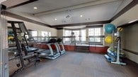 This fitness center offers a thoughtfully designed, split-level layout that organizes workout zones across two distinct areas connected by a short staircase. The lower level serves as the primary cardio and free-movement zone, featuring multiple treadmills, a ceiling fan, and recessed lighting overhead. The flooring throughout is covered in durable, tile-patterned carpet in neutral tones, providing a practical and comfortable surface for varied workouts. A large mirrored wall runs along one side, amplifying the sense of space and allowing users to monitor their form during exercise. A dedicated rack holds an assortment of stability and exercise balls in multiple sizes, adding a versatile functional training option. The upper level, accessed via the short staircase and bordered by a metal railing, houses additional cardio equipment including an elliptical and stationary bike, positioned in front of a curved bay window arrangement that fills the area with natural light and exterior views. The lower section on the opposite side contains a multi-functional cable and strength training machine. The interior color scheme features warm beige walls accented by a deep red wainscoting band and dark brown trim, contributing to a structured yet inviting athletic environment.