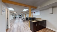 This well-appointed single-floor space is designed to serve as a versatile community or amenity room, thoughtfully divided into distinct functional zones that flow seamlessly from one end to the other. The layout is anchored by warm light wood-tone flooring that runs throughout, lending a clean and cohesive aesthetic across the entire area.

At one end, a comfortable lounge area invites relaxation, featuring a dark upholstered sofa paired with patterned accent chairs and a sleek metallic coffee table. A large decorative wall canvas adds an artistic focal point, while potted greenery introduces a refreshing natural element. Skylights above allow natural light to fill the space, enhancing its open and airy feel.

The kitchenette area serves as a practical hub, equipped with dark espresso cabinetry, a granite-topped counter, an integrated sink, and a microwave. A stylish pendant light in deep red adds a warm accent above the counter, complementing the bold yellow-painted structural columns that define zones throughout the space.

The dining area at the far end accommodates multiple tables with black stackable chairs, suitable for group gatherings or meetings. Colorful artwork adorns the walls, and flush-mounted ceiling light panels provide consistent, even illumination. Windows fitted with vertical blinds allow for natural light while maintaining privacy.