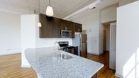 Located at 345 East Deshler Avenue in Columbus, Ohio's vibrant 43206 zip code, this single-floor space combines modern finishes with classic architectural character. The open-concept kitchen serves as a centerpiece, showcasing a generous granite island with an undermount stainless steel sink and elegant pendant lighting overhead. Dark espresso cabinetry provides ample storage and contrasts beautifully against the crisp white subway tile backsplash, while stainless steel appliances — including a range, built-in microwave, and full-size refrigerator — complete the modern aesthetic. One of the most distinctive architectural features is the ornate pressed tin ceiling, which adds a touch of historic charm and visual interest to the space. Exposed structural beams further enhance the industrial-chic character of the interior. Warm hardwood flooring flows throughout, grounding the space and adding richness to the overall design. The open layout creates a seamless flow between the kitchen and the adjoining living areas, making the space feel airy and well-connected. Large white walls offer a clean, bright backdrop that maximizes the sense of openness. This space thoughtfully blends period details with contemporary upgrades for a truly distinctive living environment.