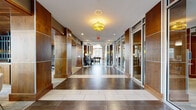 This single-story space is designed with a contemporary aesthetic, combining warm wood paneling, large-format dark tile flooring, and modern finishes throughout. The main corridor serves as an impressive entry point, lined with rich walnut-toned wall panels accented by aluminum trim and floor-to-ceiling glass partitions that allow views into adjacent rooms. Decorative drum pendant lighting punctuates the hallway, adding warmth to the polished interior.

The lounge area features soaring double-height ceilings with expansive floor-to-ceiling windows that flood the space with natural light and provide views of the surrounding landscaping. Stylish seating, a wall-mounted television, and curated décor create an inviting, well-appointed gathering space.

The kitchen is fully equipped for event use, featuring stainless steel appliances, a professional-grade range with a stainless hood, quartz countertops, warm wood cabinetry, and a textured tile backsplash. A wall of windows brings in abundant natural light and outdoor views.

A glass-enclosed sunroom offers a relaxed retreat with wraparound windows overlooking mature trees and garden beds, complemented by a generous sectional sofa and a wood-paneled ceiling that adds a natural, organic element to the space.