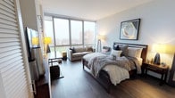This thoughtfully designed single-floor residence offers a seamless blend of modern aesthetics and functional living. The main living and sleeping area is a standout, anchored by expansive floor-to-ceiling windows that flood the space with natural light and frame panoramic views of the surrounding landscape, including green spaces and neighboring buildings. Rich, dark hardwood-style flooring runs throughout, providing a warm contrast to the crisp white walls and contemporary furnishings.

The kitchen is sleek and efficient, featuring dark cabinetry that extends to the ceiling, white quartz countertops, a white tile backsplash, and a full suite of stainless steel appliances including a gas range, dishwasher, and refrigerator. Recessed lighting and under-cabinet illumination complete the polished look.

The bathroom is finished to a high standard, offering a combination tub and shower, a dark wood vanity with a white countertop, and an LED-backlit mirror that adds a sophisticated touch. Gray hardwood-style flooring ties the aesthetic together.

Ample closet space is thoughtfully integrated into the layout, alongside in-unit laundry hookups. A cozy sitting nook positioned beside the floor-to-ceiling windows offers an inviting spot to take in the views, rounding out this well-considered and stylish urban residence.