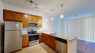This single-floor residential unit offers a thoughtfully designed layout that balances functionality and comfort throughout its living spaces. The kitchen serves as a central highlight, showcasing warm honey-toned cabinetry, granite countertops, and a full suite of stainless steel appliances including a refrigerator, electric range, built-in microwave, and dishwasher. A peninsula with a granite surface and an undermount sink provides additional workspace and defines the transition to the adjacent living area. Track lighting overhead and elegant pendant lights above the peninsula illuminate the space effectively. Light hardwood flooring runs through the kitchen and living areas, adding warmth to the clean, neutral-toned interior. The living space features large vertical-blind-covered sliding doors that allow for natural light and access to the exterior. The bedroom is carpeted and includes a window with blinds, offering a quiet and comfortable retreat. A generously sized walk-in closet with wire shelving provides ample storage. The bathroom is finished with a granite-topped vanity with an undermount sink, a large mirror, tile flooring, and a soaking tub with a tiled surround. Throughout the unit, clean white walls and consistent finishes create a cohesive, move-in-ready environment.