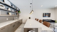 Located at 140 E Monument Ave in Dayton's historic Monument Avenue corridor, this single-floor amenity space showcases a clean, contemporary design throughout. Large-format stone-look tile flooring flows seamlessly across the entire space, grounding the modern aesthetic and lending a polished, cohesive feel.

The kitchenette area is a standout feature, outfitted with sleek gray cabinetry, white quartz countertops, and an undermount sink. Two tiers of open floating shelves line the accent wall above, providing display and storage space while adding depth and visual interest. A generous white quartz island extends into the room, accommodating several bar-height chairs for casual seating and gathering.

Adjacent to the kitchenette, a lounge area offers comfortable mixed seating arranged around a wall-mounted flat-screen television. Floor-to-ceiling glazing and a glass-panel entry door flood this zone with natural light and provide direct views of and access to an outdoor terrace, seamlessly connecting interior and exterior spaces.

A dedicated games room rounds out the layout, featuring an air hockey table, a wall-mounted television, and additional flexible seating. An open flex area with clean white walls and recessed lighting provides further versatile space. Together, these zones create a well-appointed, multi-functional environment suited for social and recreational use.