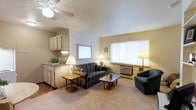 This single-floor unit features an open-concept design that seamlessly integrates the living area with a compact kitchenette. The living space is carpeted and comfortably sized, accommodating a striped sofa, a green armchair, a wooden coffee table, and a side table with a lamp, creating a welcoming and functional seating arrangement. Natural light filters in through two windows, one of which houses a wall-mounted air conditioning unit, ensuring year-round comfort. A ceiling fan overhead provides additional air circulation. The kitchenette, positioned to one side, is equipped with light wood cabinetry, a sink, and under-cabinet lighting, making it a practical space for meal preparation. The walls are painted in soft, neutral tones that contribute to the overall warm ambiance. The transition between the kitchenette's hard flooring and the carpeted living area creates a subtle but clear distinction between the two functional zones, while maintaining the open feel of the space.