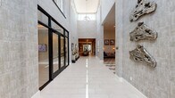 This thoughtfully designed amenity facility offers a full suite of social, recreational, and professional spaces unified by a sophisticated contemporary aesthetic. The main clubroom serves as the heart of the space, featuring a striking geometric-patterned tile floor, soaring ceilings, and a built-in wet bar with open shelving. Tufted leather sofas anchor a lounge area outfitted with wall-mounted flat-screen TVs and warm sconce lighting set against textured gray walls, creating an inviting atmosphere for gathering. Intimate booth-style seating with hairpin-leg tables and abstract artwork lining the tall walls provides additional social areas within the clubroom. A private conference room offers an elegant oval dark-wood table surrounded by plush velvet chairs with gold-toned legs, lit by a sculptural geometric pendant fixture, with clerestory windows allowing natural light. A game and lounge area includes a shuffleboard table, leather seating, and curated artwork. The dedicated business center features a row of workstations with computers positioned beneath a large window overlooking the exterior. The fitness center is generously sized with floor-to-ceiling windows, abundant natural light, and a comprehensive array of cardio and strength equipment topped by a statement oversized ceiling fan.