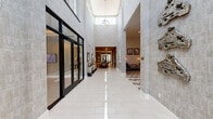 This thoughtfully designed amenity facility offers a full suite of social, recreational, and professional spaces unified by a sophisticated contemporary aesthetic. The main clubroom serves as the heart of the space, featuring a striking geometric-patterned tile floor, soaring ceilings, and a built-in wet bar with open shelving. Tufted leather sofas anchor a lounge area outfitted with wall-mounted flat-screen TVs and warm sconce lighting set against textured gray walls, creating an inviting atmosphere for gathering. Intimate booth-style seating with hairpin-leg tables and abstract artwork lining the tall walls provides additional social areas within the clubroom. A private conference room offers an elegant oval dark-wood table surrounded by plush velvet chairs with gold-toned legs, lit by a sculptural geometric pendant fixture, with clerestory windows allowing natural light. A game and lounge area includes a shuffleboard table, leather seating, and curated artwork. The dedicated business center features a row of workstations with computers positioned beneath a large window overlooking the exterior. The fitness center is generously sized with floor-to-ceiling windows, abundant natural light, and a comprehensive array of cardio and strength equipment topped by a statement oversized ceiling fan.