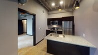 This single-floor unit at 230–298 E Grand Ave in Eau Claire, Wisconsin, blends industrial aesthetics with modern functionality in a thoughtfully designed layout. Throughout the space, polished terrazzo flooring provides a distinctive, durable foundation, while soaring ceilings with exposed ductwork and structural elements reinforce the building's urban character.

The kitchen serves as a central feature, anchored by a granite-topped island with a deep undermount sink and a contemporary arc faucet. Dark espresso cabinetry lines the walls, complemented by stainless steel appliances including a refrigerator, electric range, and built-in microwave. Pendant lighting with matte black and copper-toned fixtures illuminates both the kitchen and adjacent living areas, adding warmth to the industrial palette.

A separate room offers versatility as a bedroom or office, featuring dual black-framed windows that draw in natural light and frame views of the surrounding Eau Claire cityscape. A bold dark-panel door anchors the space with a sleek, modern touch.

The bathroom continues the cohesive design language with granite countertops, a dark wood vanity, an undermount sink, and terrazzo flooring. Neutral gray walls throughout the unit create a calm, cohesive backdrop that ties each space together seamlessly.